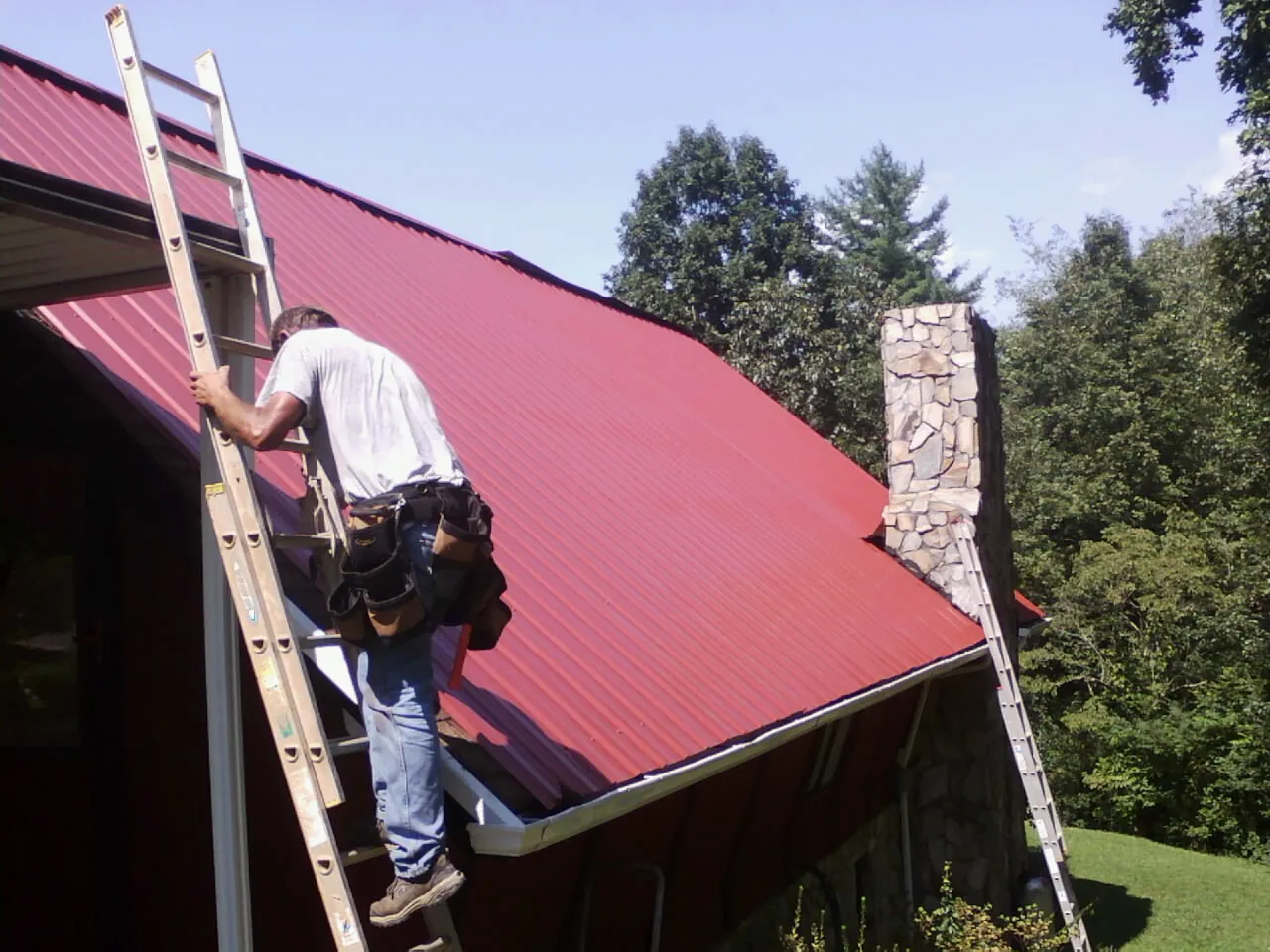Roofer on a ladder beside a red metal roof and stone chimney — Soffit & Fascia Repair by Roofing Brothers in Vernon